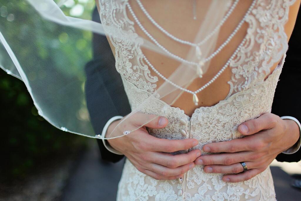Braeutigam umarmt laechelnde Braut im weissen Spitzenkleid mit Perlenkette