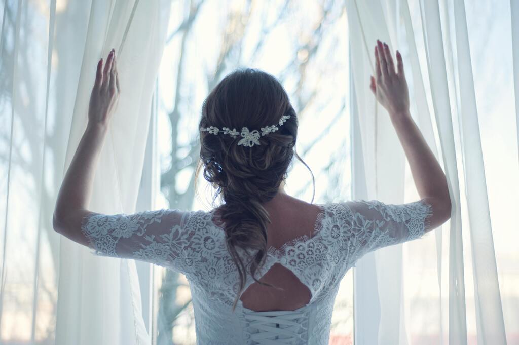 Braut in weissem Spitzenkleid mit Blumenhaarschmuck am Fenster, ruhig